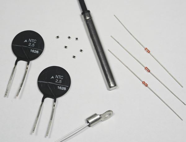 Photograph showing multiple thermistor types, including disc-shaped NTC thermistors, bead thermistors with wire leads, small surface‑mount components, and temperature probes arranged on a white surface.