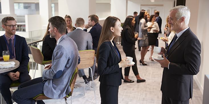 A group of people in business attire standing and sitting in a bright office lounge, talking with one another while holding coffee cups and light refreshments during a professional networking break.
