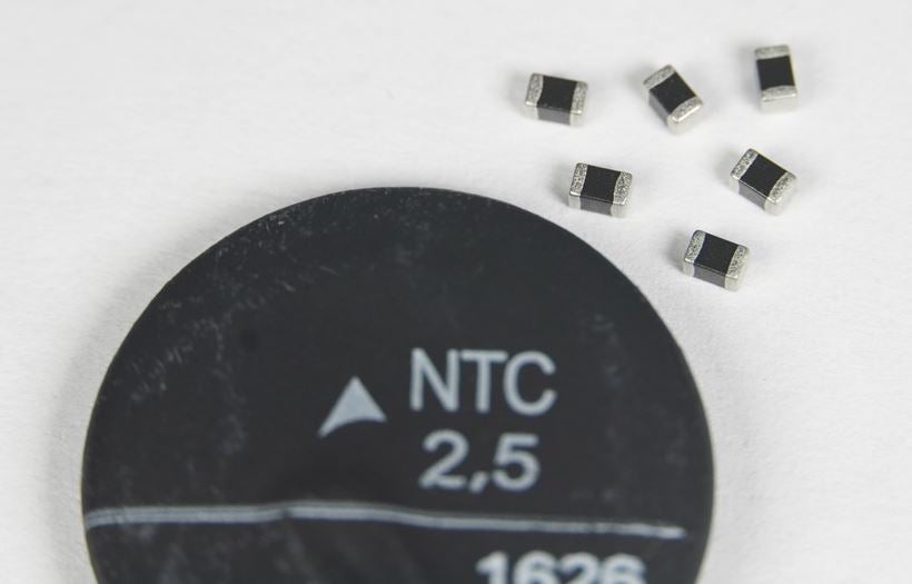 Close-up photograph of an NTC thermistor disc labeled ‘NTC 2.5’ with several small surface‑mount electronic components on a white background.