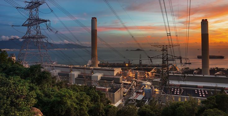 Gran instalación de energía eléctrica con torres de transmisión y líneas de alta tensión cerca de una planta industrial al atardecer.