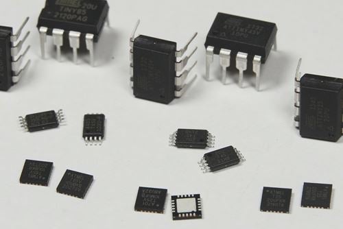 Various electronic integrated circuits in different package sizes, including DIP and sComparison of two Atmel microcontroller chips, one larger DIP package standing on metal pins and one smaller surface‑mount package, shown on a white background.