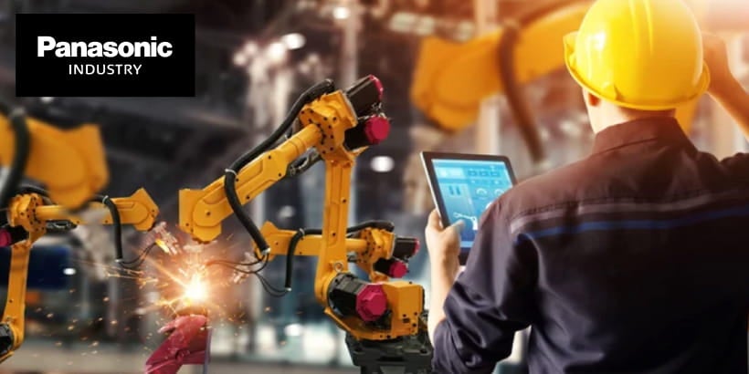 Une personne portant un casque jaune utilise une tablette tout en surveillant les bras robotiques de Panasonic Industry dans un environnement industriel.