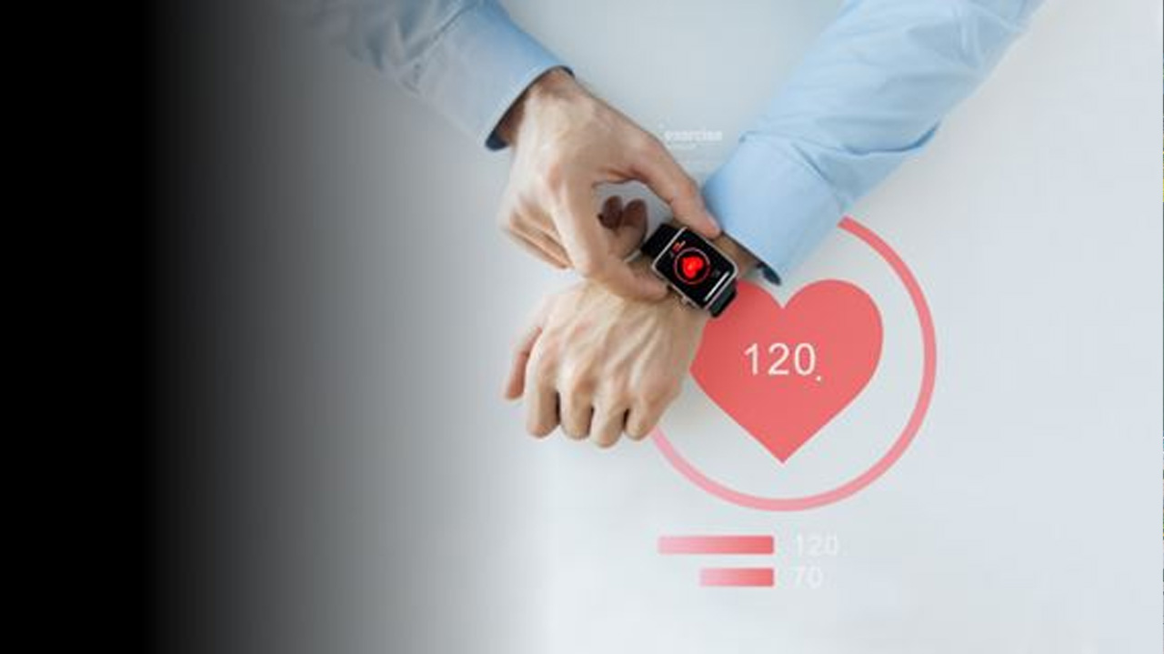 A man in a light blue shirt interacts with a smartwatch on his wrist, which displays a heart icon and a digital heart rate reading. 