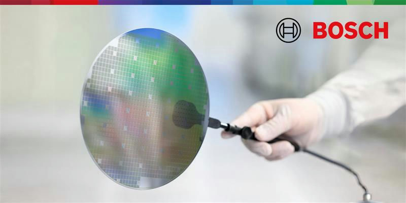A gloved hand holds a reflective semiconductor silicon wafer with a grid of tiny squares visible on its surface. 