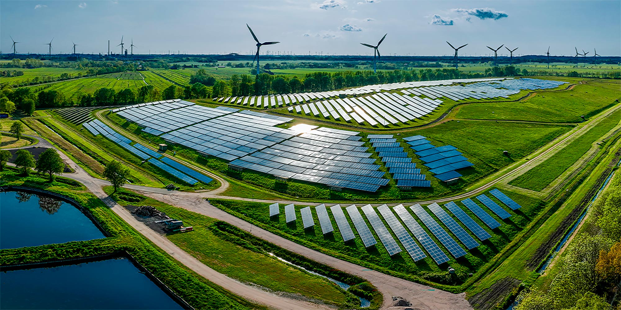 A vast solar farm stretches across green fields, with rows of reflective solar panels arranged in neat geometric patterns. 