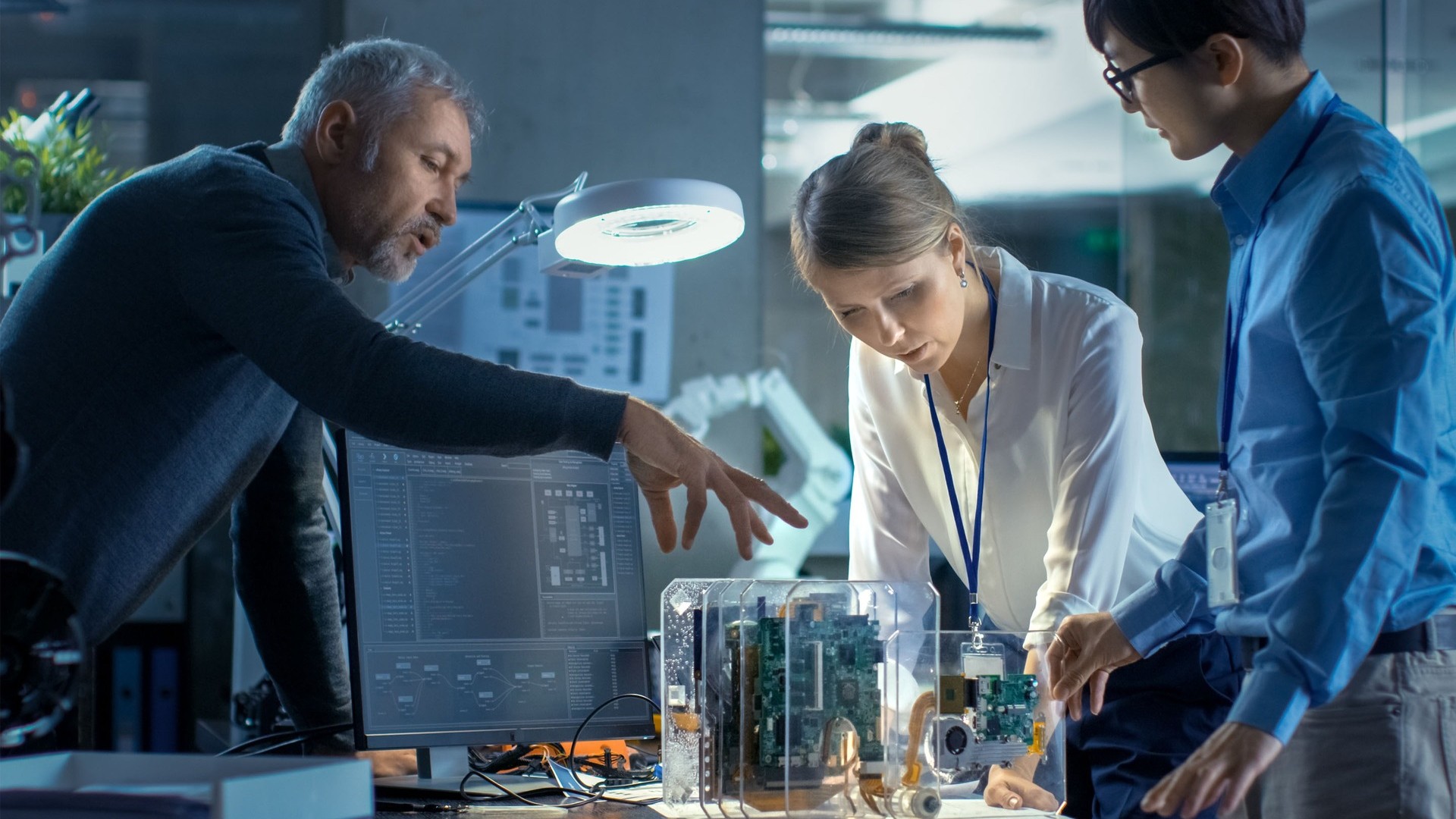 A group of professionals is working together in a modern office setting, focusing on an electronic prototype encased in a transparent housing