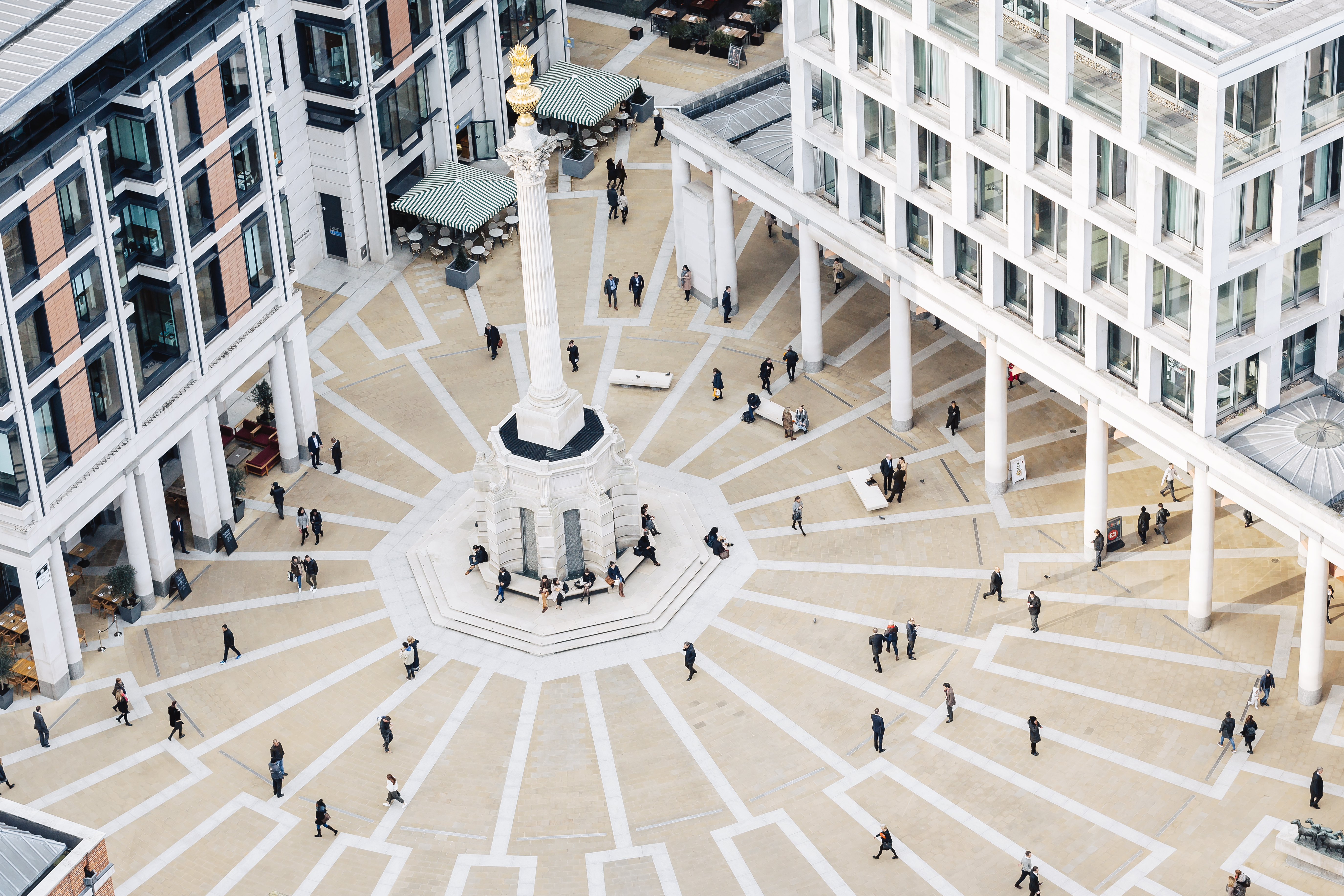 Vue aérienne de Paternoster Square à Londres, montrant la colonne centrale, les motifs rayonnants du pavage, les bâtiments modernes environnants, ainsi que des personnes marchant ou se rassemblant dans l'espace public.